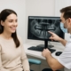 A smiling woman in a dental chair is consulting with a male dental implantologist. The doctor is pointing to a digital scan of the patient's jaw on a computer screen, illustrating the planned dental implant procedure. No text on the image.