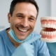Close up image of a dentist smiling and pointing to a set of perfect, white, false teeth in a patient's mouth. No text on image.