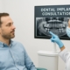 A person sitting in a dentist's chair getting a consultation about dental implants. The dentist is pointing to a scan of the patient's mouth, with the words "Dental Implant Consultation" on the screen.