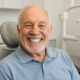 A smiling senior man is showing off his new set of All-on-4 dental implants. He is sitting in a modern dental office in Hialeah, FL. No text on image.