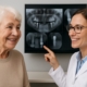A smiling senior woman is talking with a friendly, professional, middle-aged female implant dentist. The dentist is pointing to digital X-rays on a monitor in the background. No text on image.