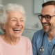 Image of a smiling elderly woman with full, bright white dentures, laughing with a dentist in a modern office to showcase full mouth dental implants. No text on image.
